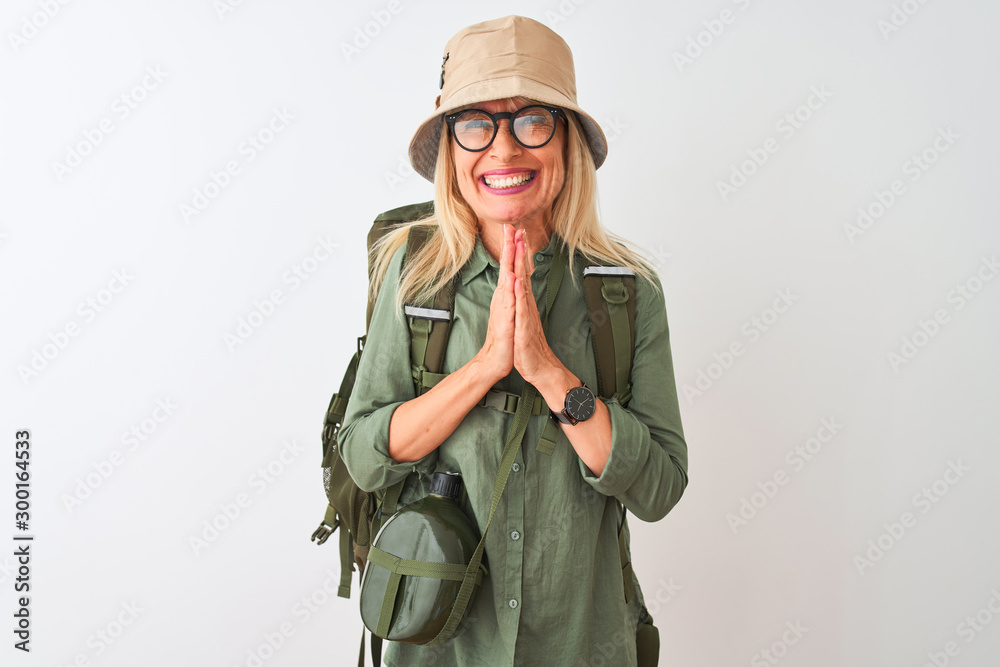 Middle age hiker woman wearing backpack hat canteen glasses over isolated white background praying with hands together asking for forgiveness smiling confident. Middle age hiker woman wearing backpack hat canteen glasses over isolated white background praying with hands together asking for forgiveness smiling confident.
