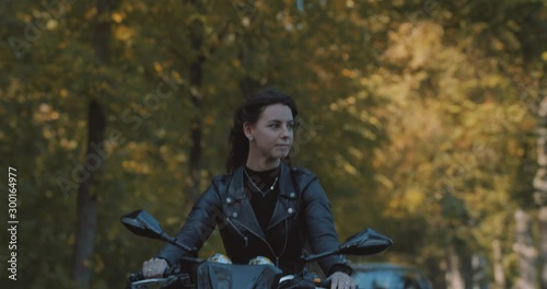 Pretty smiling European young woman driving a motorbike wearing leather jacket in forest with vibrant, colorful golden autumn leaves on sunny day. Frontal view, Slow Motion, Utrecht , Netherlands photo