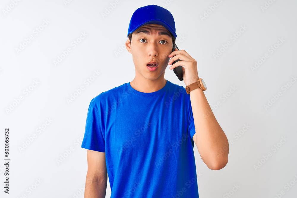 Chinese deliveryman wearing cap talking on the smartphone over isolated white background scared in shock with a surprise face, afraid and excited with fear expression