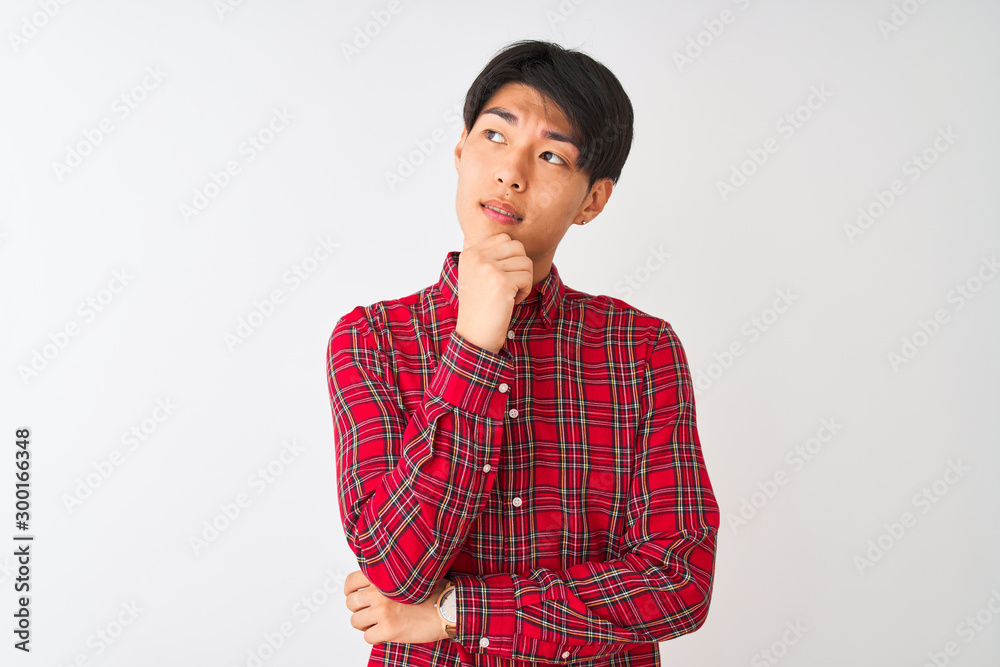 Young chinese man wearing casual red shirt standing over isolated white background with hand on chin thinking about question, pensive expression. Smiling with thoughtful face. Doubt concept.
