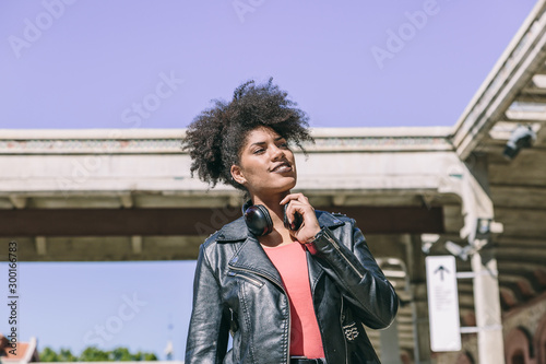 Beautiful afro haired woman with black jacket and black headphones around her neck smiles looking aside while taking a walk 