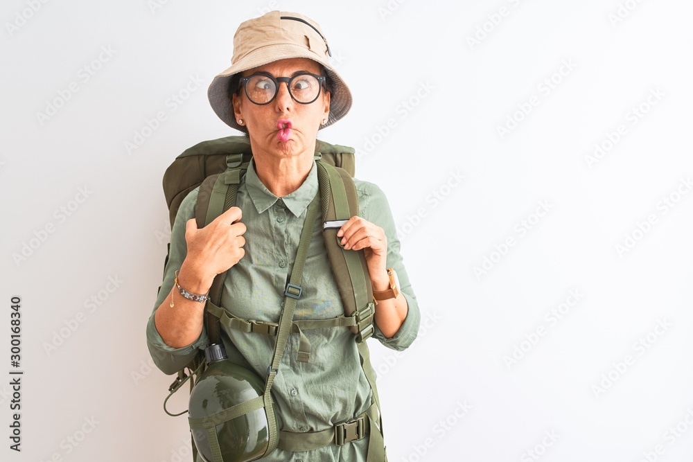 Fototapeta premium Middle age hiker woman wearing backpack canteen hat glasses over isolated white background making fish face with lips, crazy and comical gesture. Funny expression.
