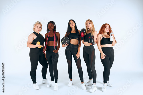 Sportive girls in sport wearing stand together with boxing gloves, dumbbells and other sportive things, look at camera isolated, white background