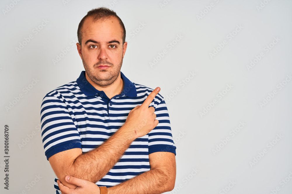 Young man wearing casual striped polo standing over isolated white background Pointing with hand finger to the side showing advertisement, serious and calm face Young man wearing casual striped polo standing over isolated white background Pointing with hand finger to the side showing advertisement, serious and calm face