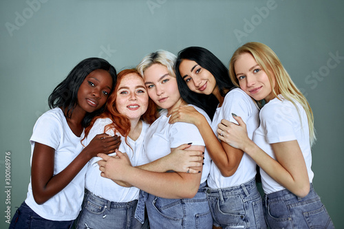 Portrait of five smiling beautiful women in jeans standing together isolated over grey background, hug each other, laugh