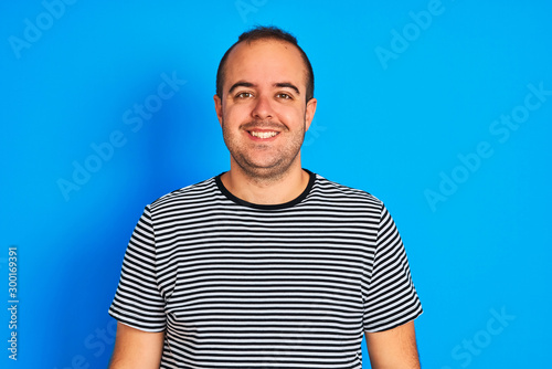 Young man wearing striped navy t-shirt standing over isolated blue background with a happy and cool smile on face. Lucky person.