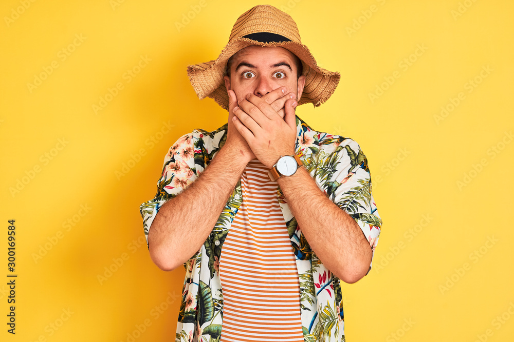 Young man on vacation wearing summer shirt and hat over isolated yellow background shocked covering mouth with hands for mistake. Secret concept. Young man on vacation wearing summer shirt and hat over isolated yellow background shocked covering mouth with hands for mistake. Secret concept.