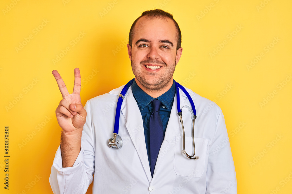 Young doctor man wearing coat and stethoscope standing over isolated yellow background showing and pointing up with fingers number two while smiling confident and happy. Young doctor man wearing coat and stethoscope standing over isolated yellow background showing and pointing up with fingers number two while smiling confident and happy.