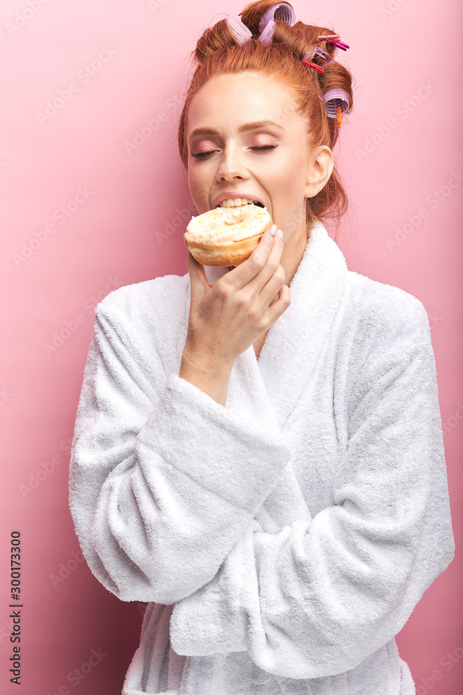 Portrait of beautiful girl with sweet tasty donut, eating after shower, dressed in white bathrobe, curlers on hair. Isolated pink background Portrait of beautiful girl with sweet tasty donut, eating after shower, dressed in white bathrobe, curlers on hair. Isolated pink background