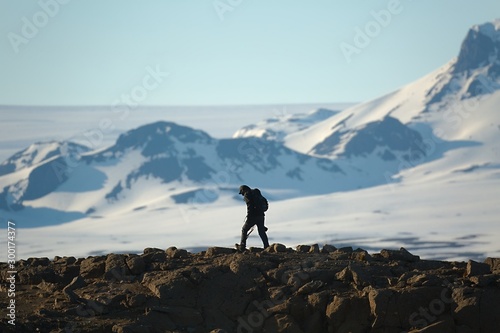 Walking on cliffs in Icelandic Highlands, snowy mountains in the background, hooded man photographer carriing a camera © Gudellaphoto