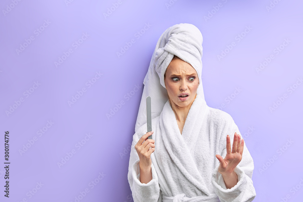 Young beautiful woman in bathrobe and towel on head look with fear on nails, cuts nails with nailfile. Isolated over purple background Young beautiful woman in bathrobe and towel on head look with fear on nails, cuts nails with nailfile. Isolated over purple background