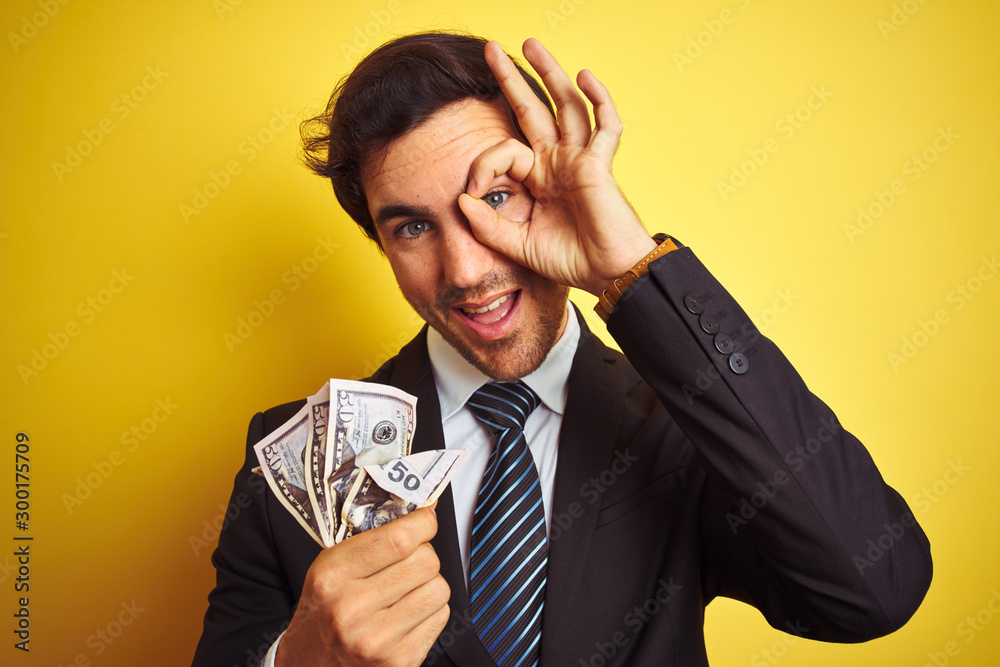 Young handsome businessman wearing suit holding dollars over isolated yellow background with happy face smiling doing ok sign with hand on eye looking through fingers Young handsome businessman wearing suit holding dollars over isolated yellow background with happy face smiling doing ok sign with hand on eye looking through fingers