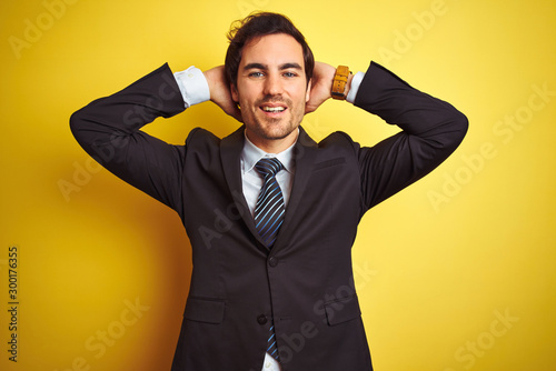 Young handsome businessman wearing suit and tie standing over isolated yellow background relaxing and stretching, arms and hands behind head and neck smiling happy