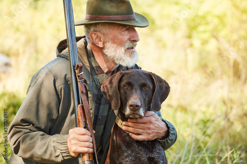 Caucasian mature man with gun and dog sit searching prey. Bearded man in hunting clothes. Autumn photo