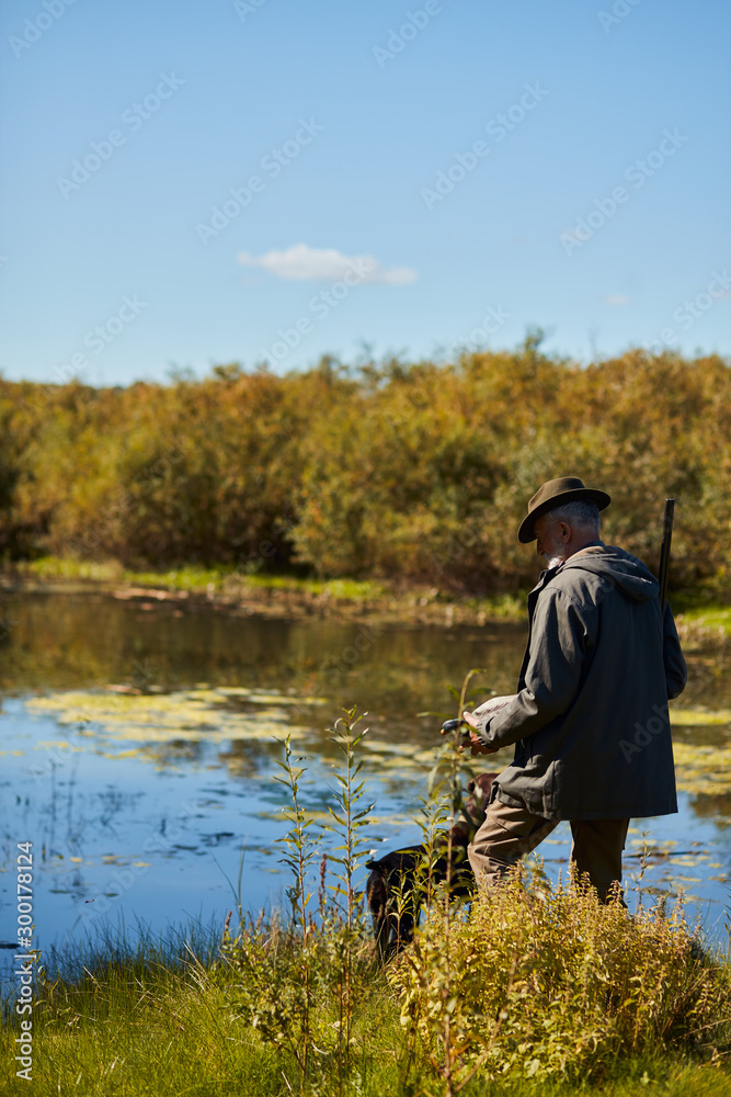 Mature man hunter in hat stand near lake with shotgun, going to hunt on wild ducks. Autumn hunting