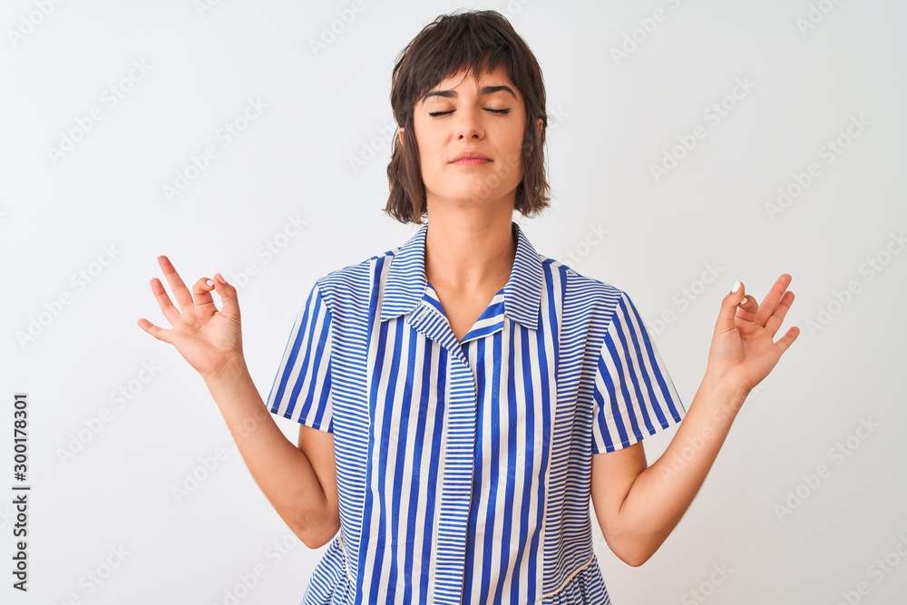 Young beautiful woman wearing blue striped shirt standing over isolated white background relax and smiling with eyes closed doing meditation gesture with fingers. Yoga concept. Young beautiful woman wearing blue striped shirt standing over isolated white background relax and smiling with eyes closed doing meditation gesture with fingers. Yoga concept.