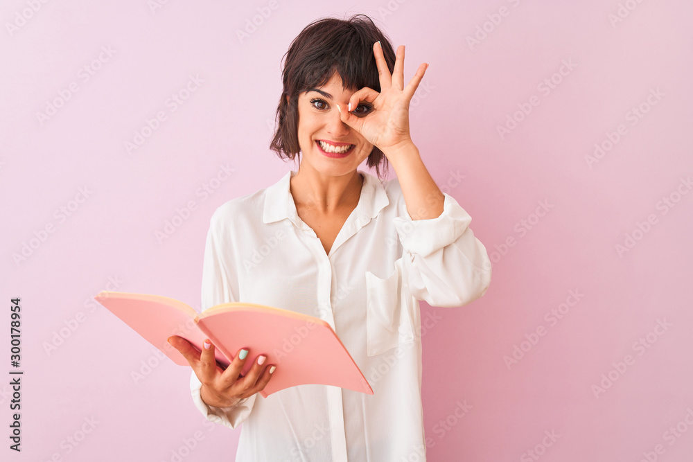 Young beautiful teacher woman reading book standing over isolated pink background with happy face smiling doing ok sign with hand on eye looking through fingers Young beautiful teacher woman reading book standing over isolated pink background with happy face smiling doing ok sign with hand on eye looking through fingers