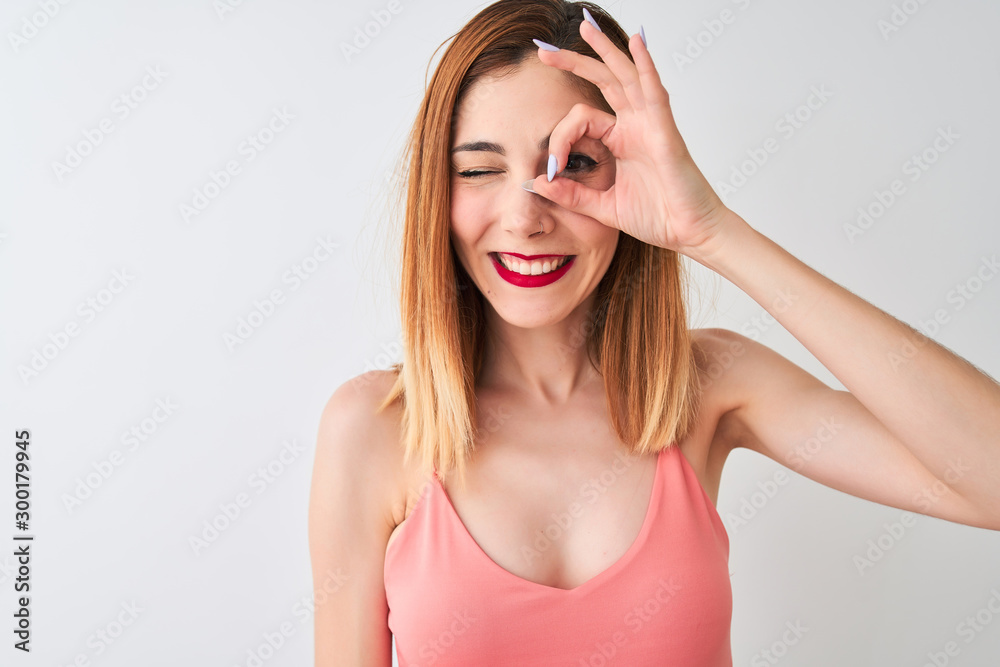 Beautiful redhead woman wearing casual pink t-shirt standing over isolated white background with happy face smiling doing ok sign with hand on eye looking through fingers