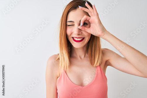 Beautiful redhead woman wearing casual pink t-shirt standing over isolated white background with happy face smiling doing ok sign with hand on eye looking through fingers