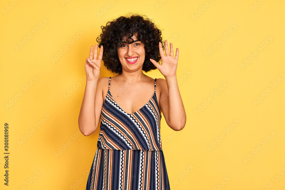 Arab woman with curly hair wearing striped colorful dress over isolated yellow background showing and pointing up with fingers number eight while smiling confident and happy. Arab woman with curly hair wearing striped colorful dress over isolated yellow background showing and pointing up with fingers number eight while smiling confident and happy.