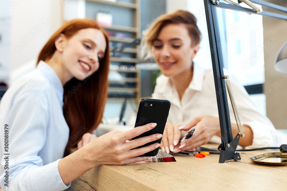 Cheerful master and client look at phone on beautiful manicure. Redhead girl want manicur like on smartphone. Cheerful master and client look at phone on beautiful manicure. Redhead girl want manicur like on smartphone.