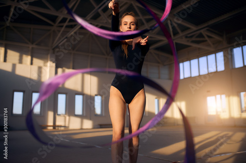 se up of young sporty woman dressed in black leotard, str tching hand towards camera, looks straight with calm face, waving bright coloured ribbon in the air, rhythmic gymnastics concept photo