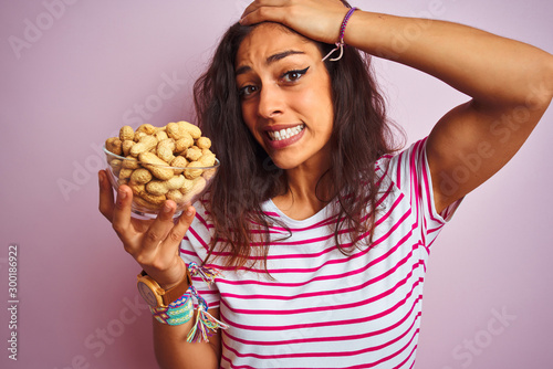 Young beautiful woman holding bowl with peanuts over isolated pink background stressed with hand on head  shocked with shame and surprise face  angry and frustrated. Fear and upset for mistake.