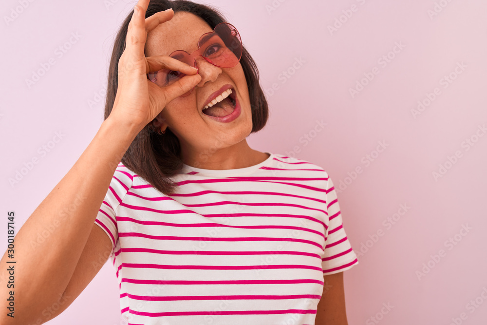 Young beautiful woman wearing fashion sunglasses with hearts over isolated pink background with happy face smiling doing ok sign with hand on eye looking through fingers Young beautiful woman wearing fashion sunglasses with hearts over isolated pink background with happy face smiling doing ok sign with hand on eye looking through fingers