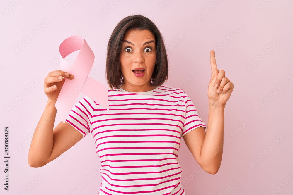Young beautiful woman holding cancer ribbon standing over isolated pink background surprised with an idea or question pointing finger with happy face, number one Young beautiful woman holding cancer ribbon standing over isolated pink background surprised with an idea or question pointing finger with happy face, number one