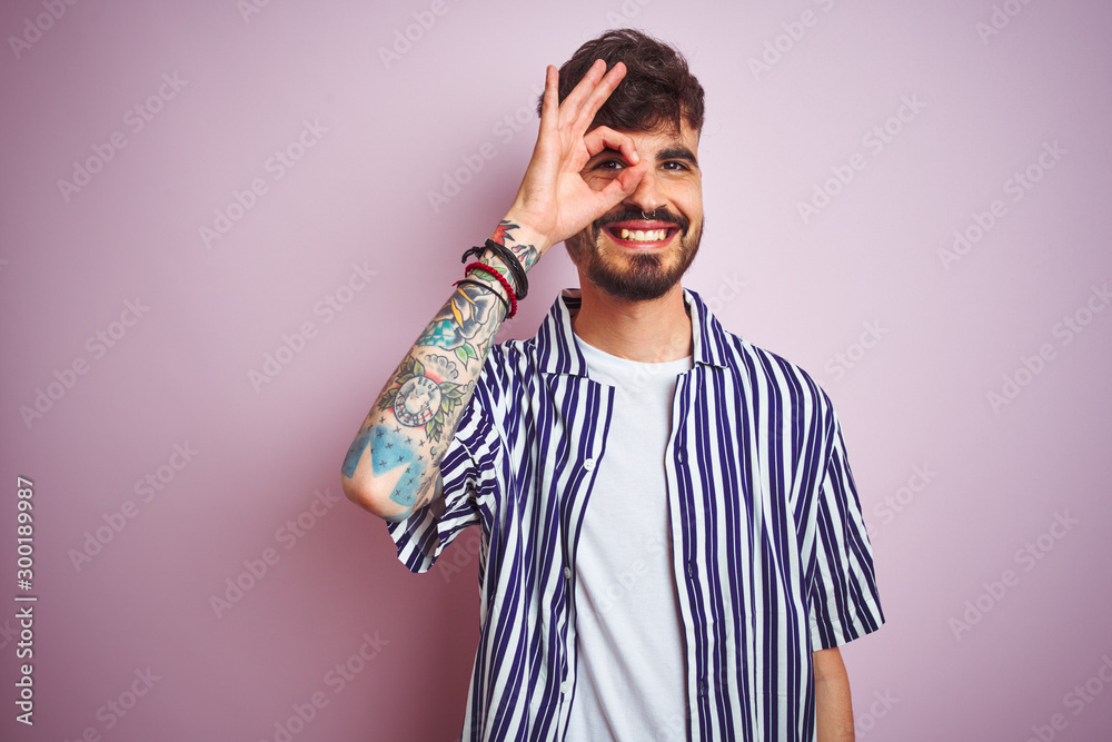 Young man with tattoo wearing striped shirt standing over isolated pink background doing ok gesture with hand smiling, eye looking through fingers with happy face. Young man with tattoo wearing striped shirt standing over isolated pink background doing ok gesture with hand smiling, eye looking through fingers with happy face.