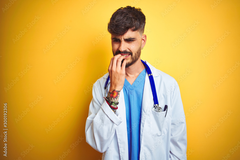 Young doctor man with tattoo wearing stethocope standing over isolated yellow background looking stressed and nervous with hands on mouth biting nails. Anxiety problem. Young doctor man with tattoo wearing stethocope standing over isolated yellow background looking stressed and nervous with hands on mouth biting nails. Anxiety problem.