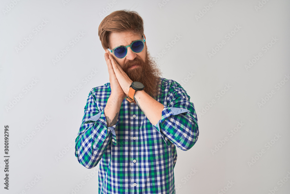 Young redhead irish man wearing casual shirt and sunglasses over isolated white background sleeping tired dreaming and posing with hands together while smiling with closed eyes. Young redhead irish man wearing casual shirt and sunglasses over isolated white background sleeping tired dreaming and posing with hands together while smiling with closed eyes.