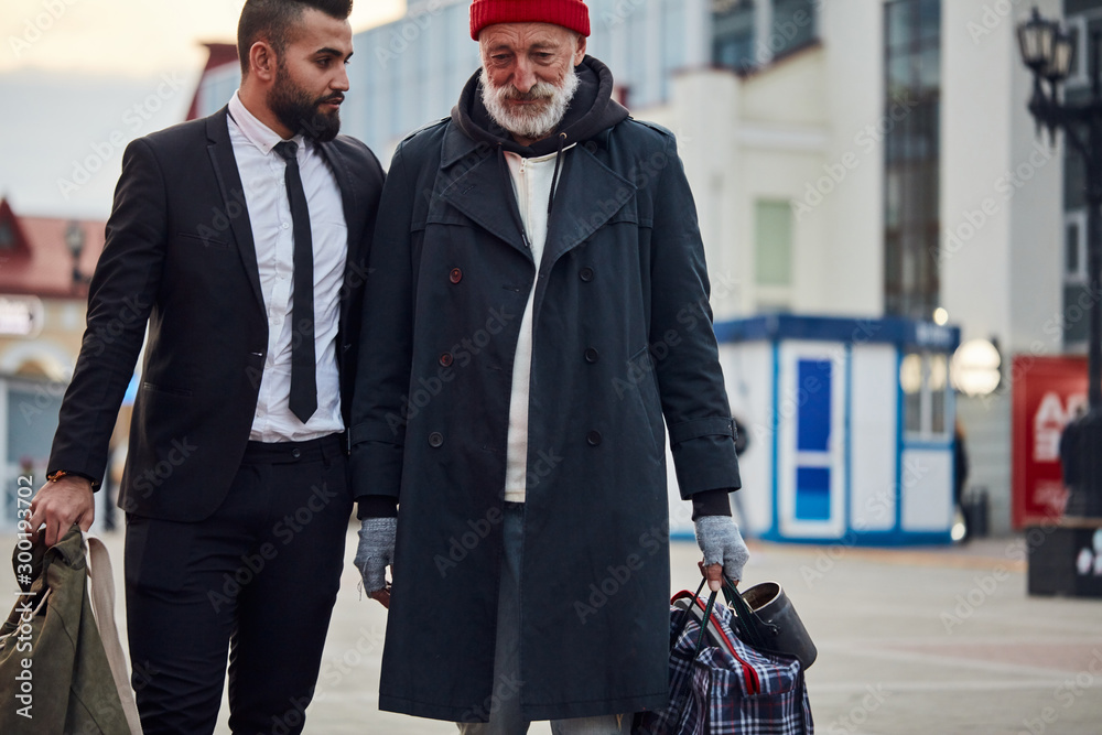 Rich man decided to help poor man. Young bearded man in black suit with beggar senior male. Contrast, different segments of society Stock Photo | Adobe Stock