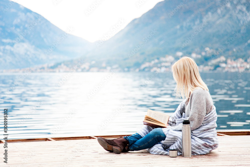 Fototapeta premium Woman is reading book by winter sea, mountains, beach. Cozy picnic on wooden pier with coffee, hot beverages, tea or cocoa in thermos, warm plaid. Girl is enjoying life, relaxation, wellbeing.