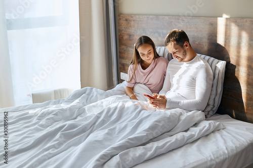 Young husband and wife having honeymoon, texting to friends about arrival to other country, travelling together, resting in hotel room, using cell phone to message, love concept