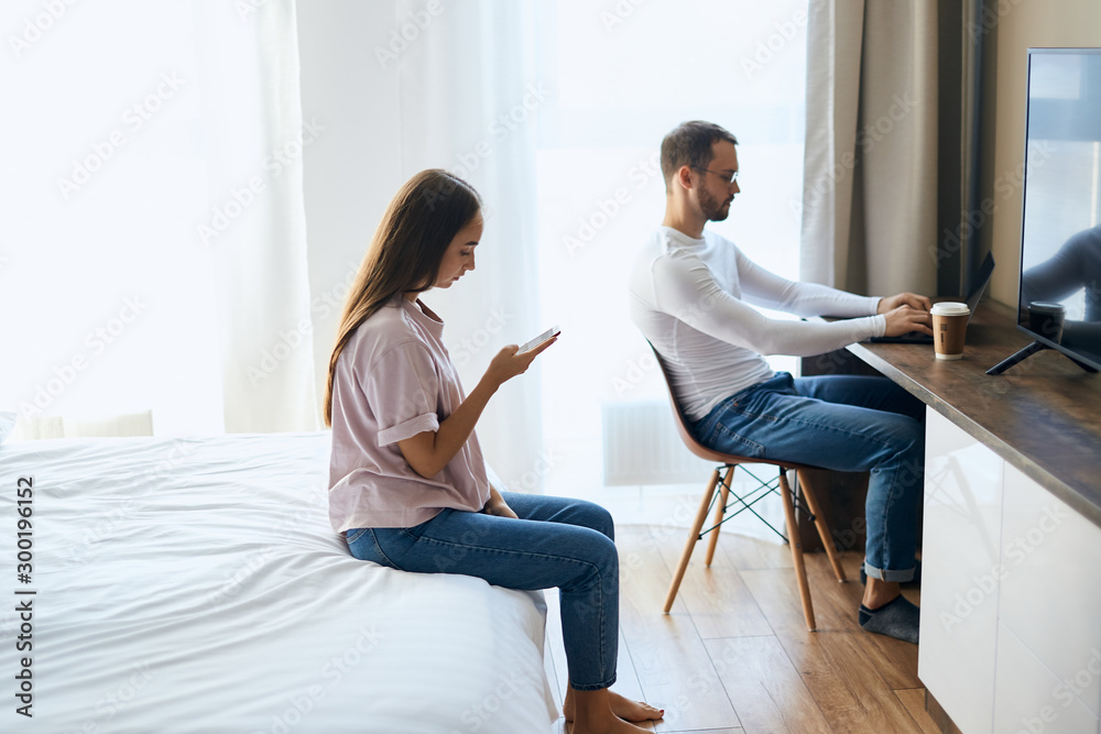 Tall young boyfriend sitting in front of laptop computer, working hard with calm face, pretty girlfriend chatting with friends using phone behind serious man, indoor shot, background of white curtains Tall young boyfriend sitting in front of laptop computer, working hard with calm face, pretty girlfriend chatting with friends using phone behind serious man, indoor shot, background of white curtains