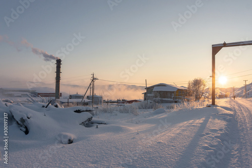 the bright orange sun at dawn in the north beyond the Arctic Circle illuminates the small grass, pipes and old rural houses on a frosty morning in winter