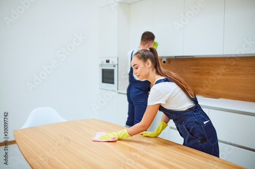 Young professionals of cleaning service do spring-cleaning in kitchen. Woman in special equipment wiping dust off from table, background man wiping dust off from cupboard