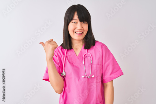 Young beautiful Chinese nurse woman wearing stethoscope over isolated white background smiling with happy face looking and pointing to the side with thumb up.