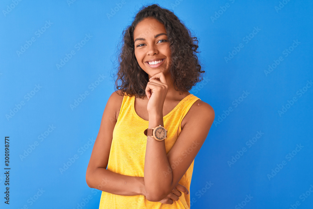Young brazilian woman wearing yellow t-shirt standing over isolated blue background looking confident at the camera smiling with crossed arms and hand raised on chin. Thinking positive. Young brazilian woman wearing yellow t-shirt standing over isolated blue background looking confident at the camera smiling with crossed arms and hand raised on chin. Thinking positive.