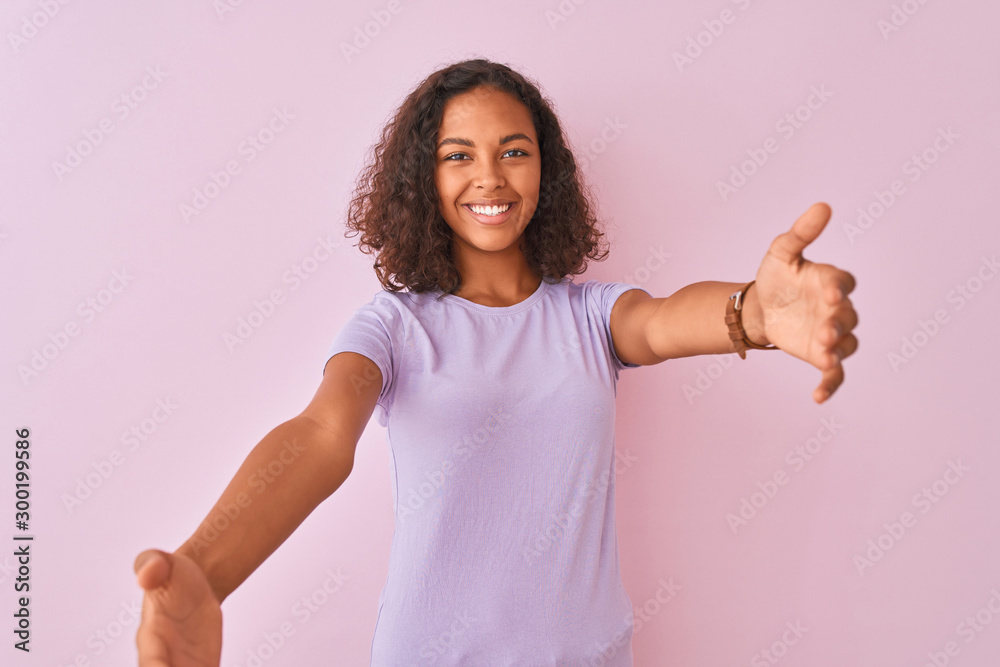 Young brazilian woman wearing t-shirt standing over isolated pink background looking at the camera smiling with open arms for hug. Cheerful expression embracing happiness. Young brazilian woman wearing t-shirt standing over isolated pink background looking at the camera smiling with open arms for hug. Cheerful expression embracing happiness.