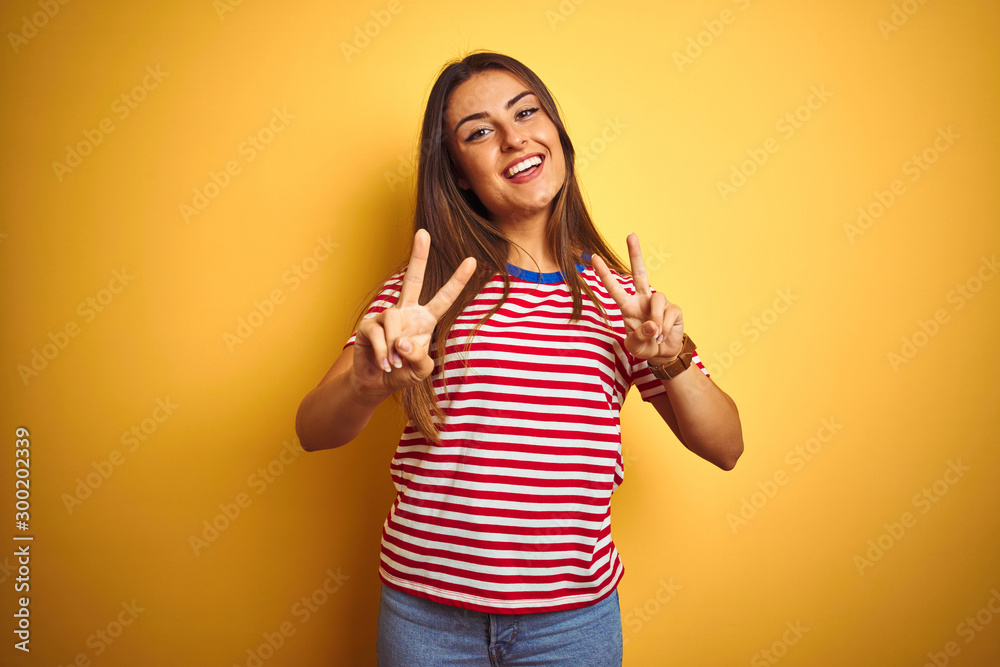 Young beautiful woman wearing striped t-shirt standing over isolated yellow background smiling looking to the camera showing fingers doing victory sign. Number two. Young beautiful woman wearing striped t-shirt standing over isolated yellow background smiling looking to the camera showing fingers doing victory sign. Number two.