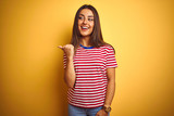 Young beautiful woman wearing striped t-shirt standing over isolated yellow background smiling with happy face looking and pointing to the side with thumb up.