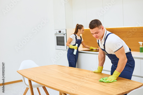 Two young cleaners in uniform effectively and quickly work together in kitchen. Youn caucasian man wiping kitchen table  woman wiping sensory electric stove