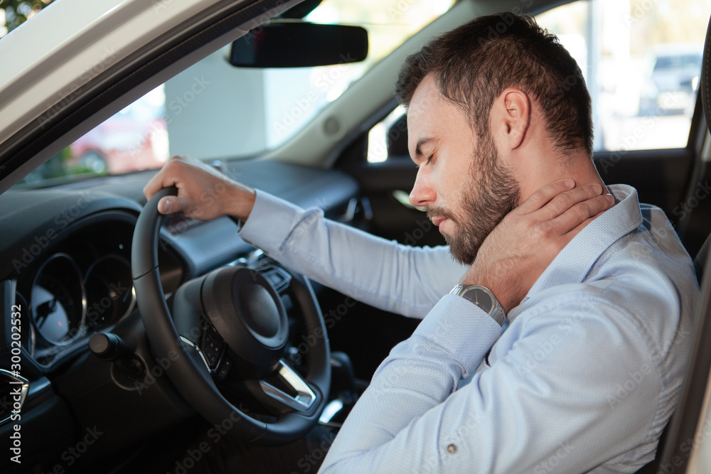 Young man rubbing his aching neck, looking tired from driving. Male driver having neck pain, sitting in his car Young man rubbing his aching neck, looking tired from driving. Male driver having neck pain, sitting in his car