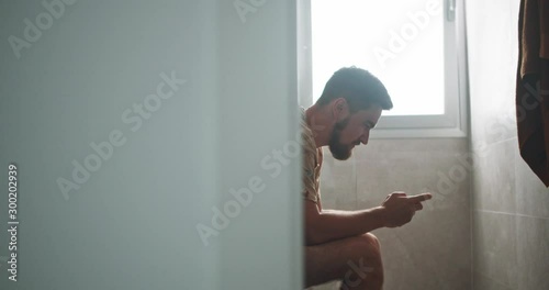 In the bathroom young man on toilet relaxing playing mobile game on smartphone winning cheering up. Fun time.