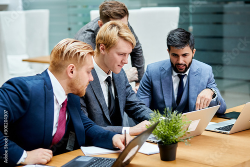 Young co-workers sitting on table and discuss project on laptop. Caucasian businessmen gathered on tabe, using modern technologies