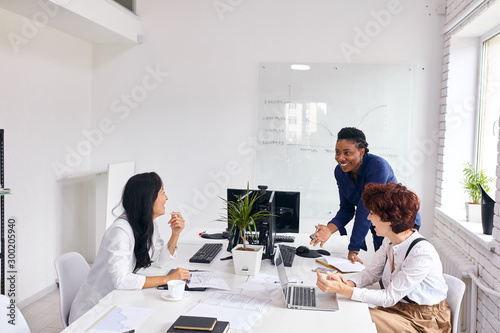 Black business coach representing business plan to caucasian ladies in office, white interior