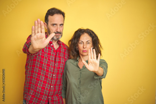 Beautiful middle age couple over isolated yellow background doing stop sing with palm of the hand. Warning expression with negative and serious gesture on the face.