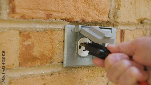 Man plugging in a power cord and then unplugging it from an outdoor electrical outlet. Dual covered outdoor power sockets on brick wall. Orange extension cord plug inserted into a wall socket.